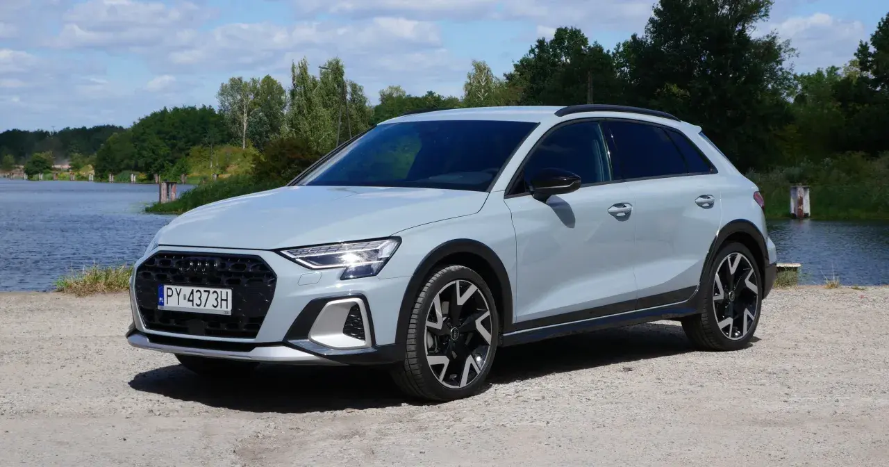 Jasnoszary Audi A3 Allstreet z czarnymi dodatkami stoi na żwirowej drodze nad jeziorem.