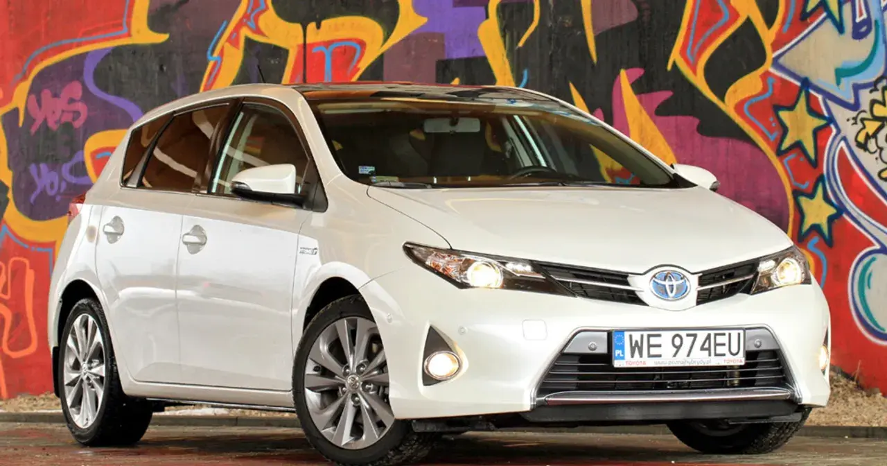 Biała Toyota Auris 2015 z polskimi tablicami rejestracyjnymi WE 974EU zaparkowana przed kolorowym muralem.