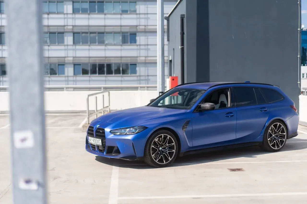 Niebieskie BMW G81 na parkingu, z nowoczesnym budynkiem w tle.