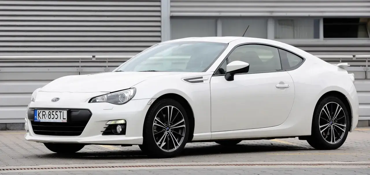 Białe Toyota BRZ z czarnymi felgami stoi przed budynkiem z poziomymi żaluzjami.