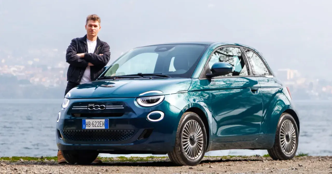 Młody mężczyzna stoi obok swojego nowego, turkusowego fiat mini 500 nad jeziorem.