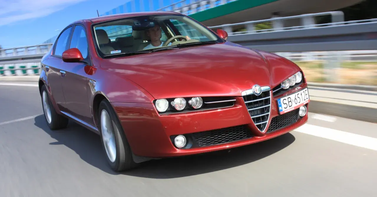 Czerwone Alfa Romeo 159 1.9 JTD pędzi po drodze, kierowca skupiony na trasie.