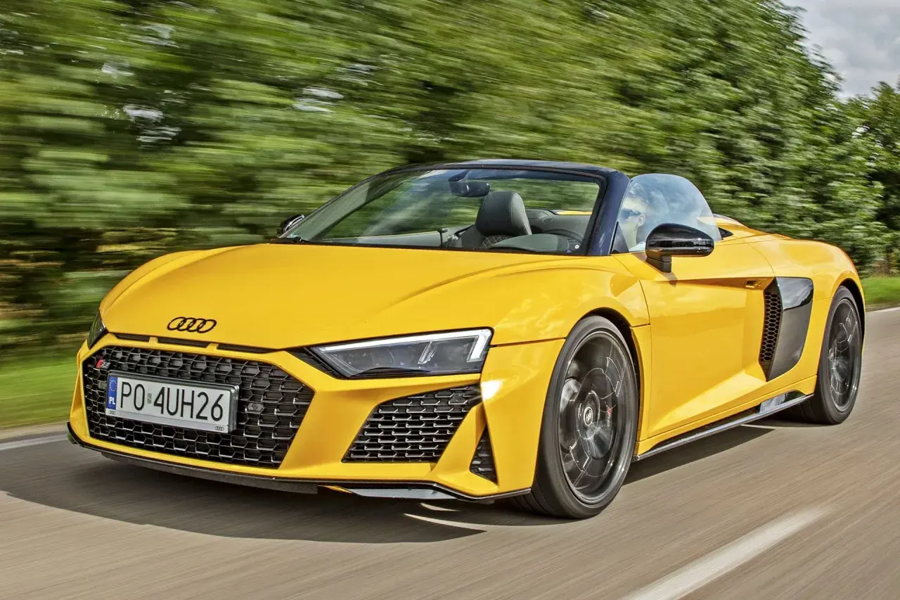 Żółty kabriolet Audi R8 V10 pędzi po drodze, otoczony rozmytymi zielonymi drzewami.