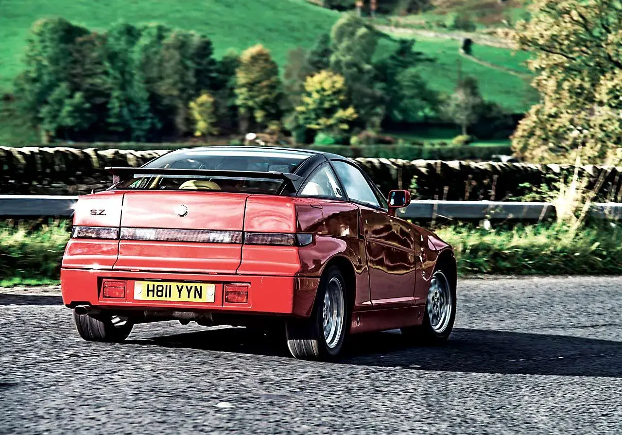 Czerwone Alfa Romeo SZ z czarnym dachem i spoilerem jedzie po wiejskiej drodze.