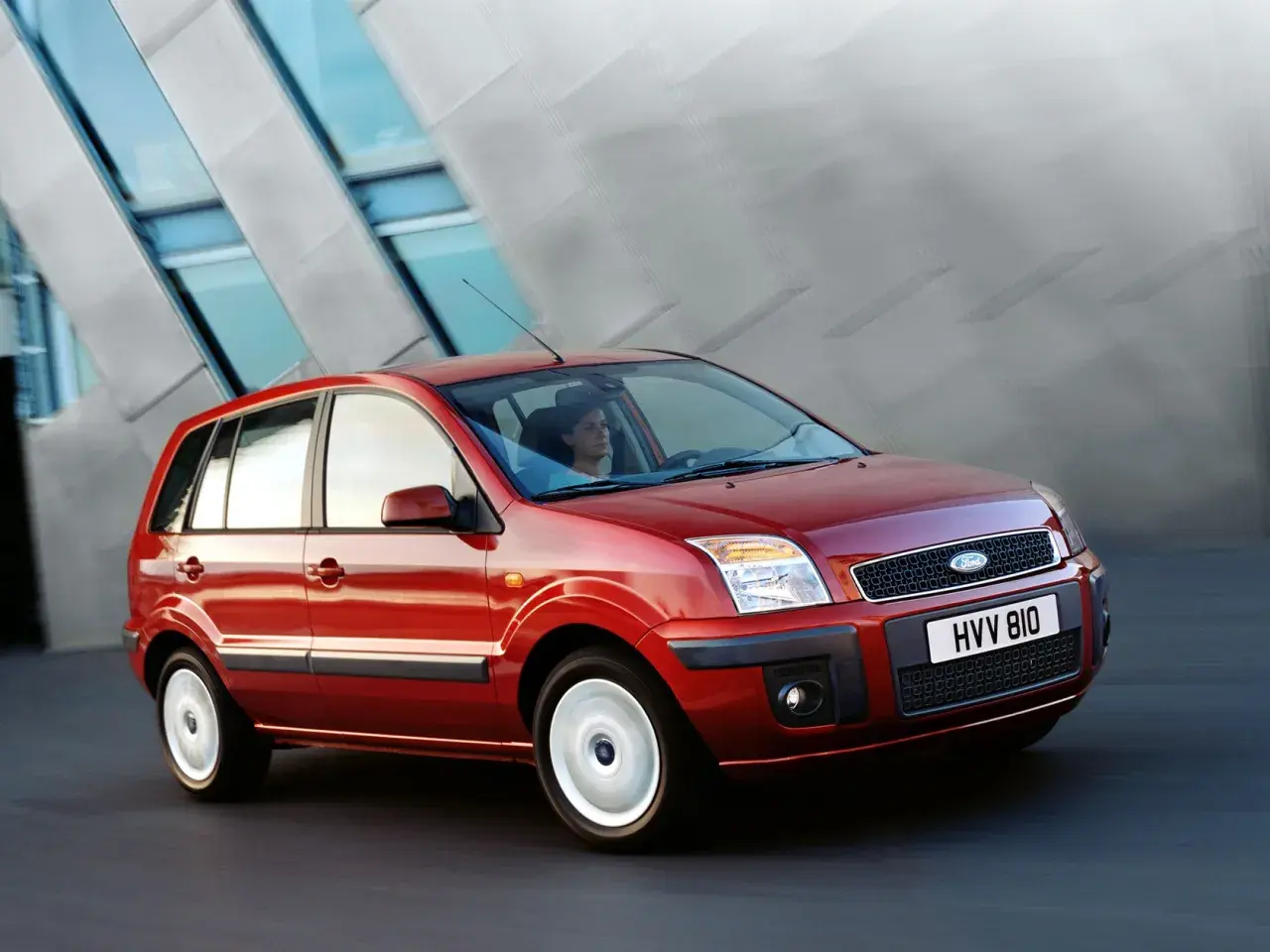 Czerwony Ford Fusion 1.4 benzyna w ruchu, z kobietą za kierownicą, na tle nowoczesnego budynku.