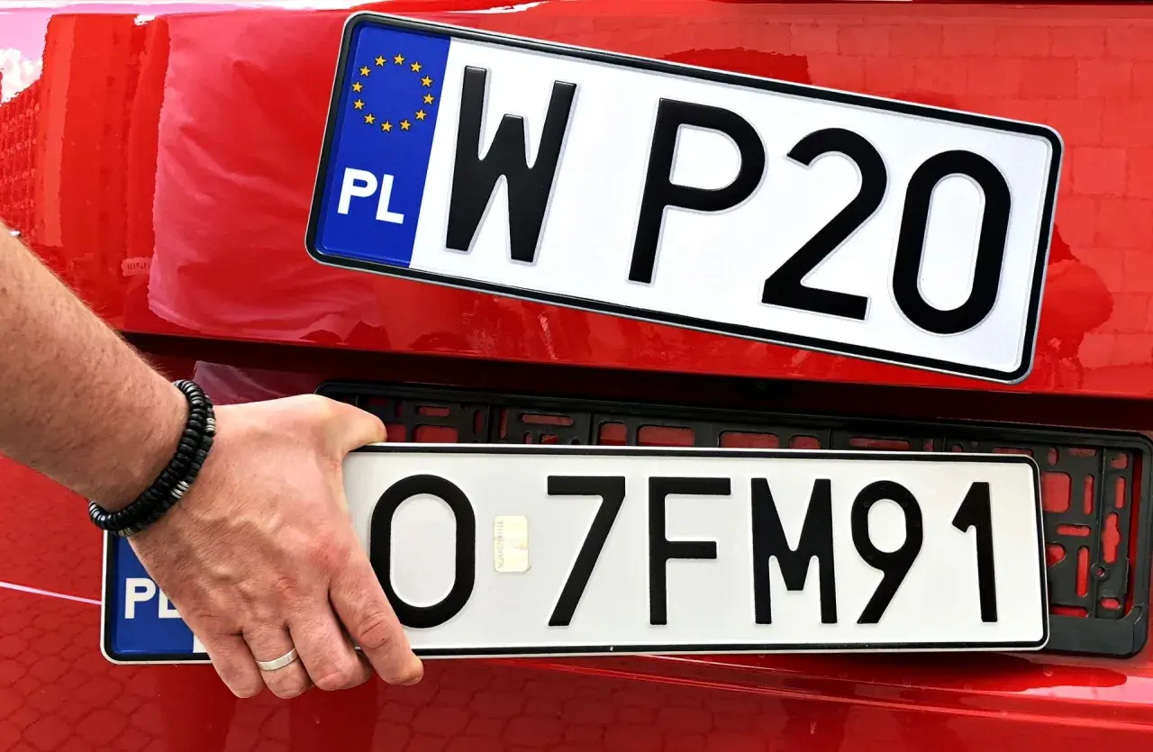 Ręka z bransoletką montuje tablicę rejestracyjną z napisem "O 7FM91" na czerwonym samochodzie. Powyżej widać inną tablicę "W P20".