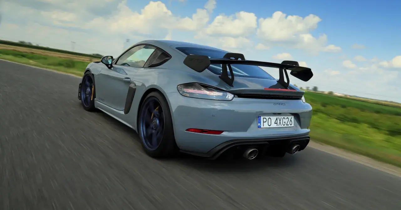 Szary Porsche GT4 RS z dużym skrzydłem pędzi po drodze, zostawiając za sobą rozmyte pola i błękitne niebo.