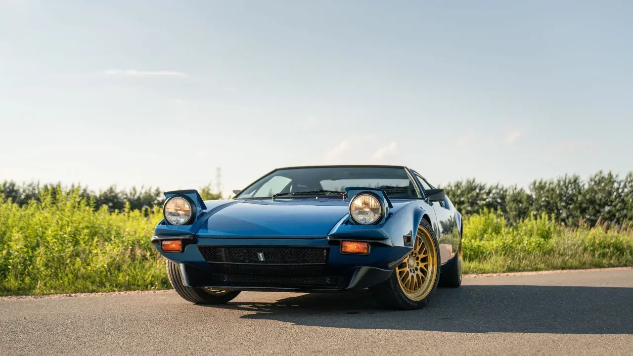 Niebieski De Tomaso Pantera z wyskakującymi reflektorami i złotymi felgami na tle zieleni i błękitnego nieba.