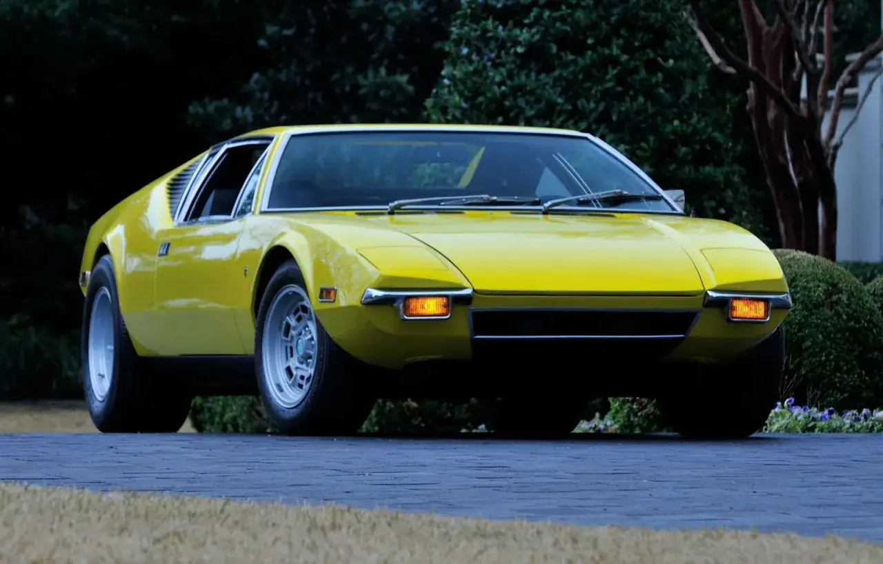 Żółty De Tomaso Pantera z lat 70. na podjeździe, otoczony zielenią.