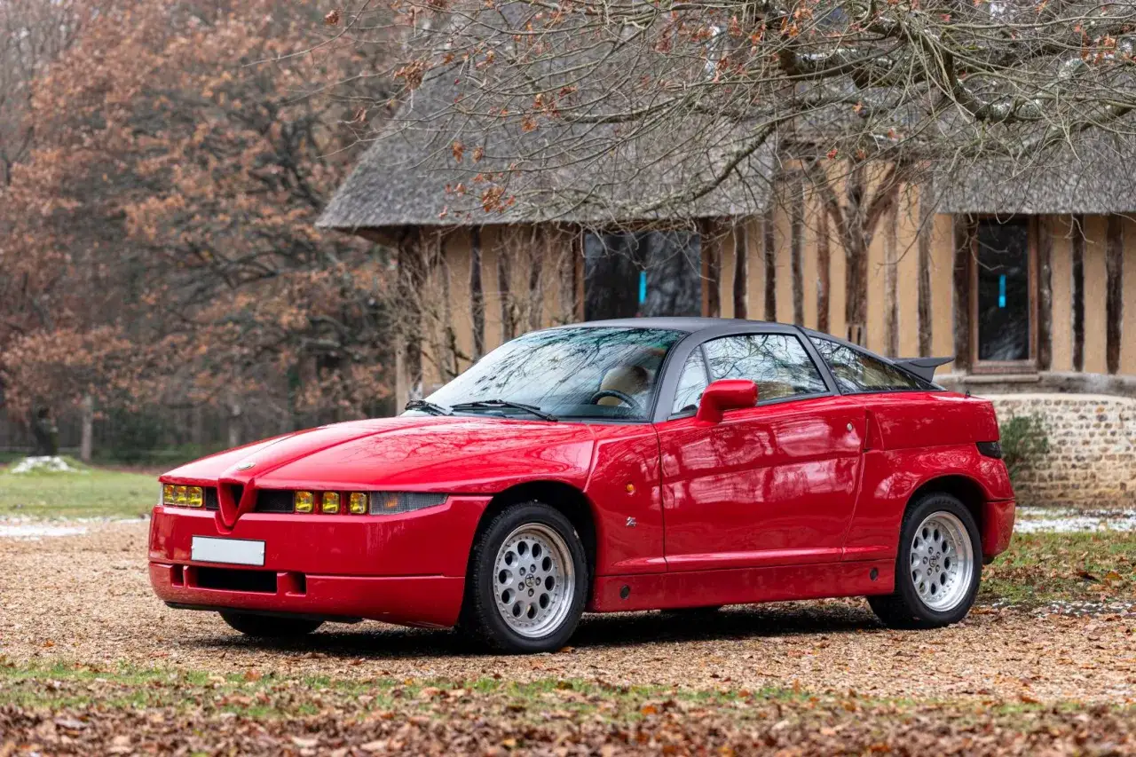Czerwone Alfa Romeo SZ z czarnym dachem i spojlerem, zaparkowane przed drewnianym budynkiem.