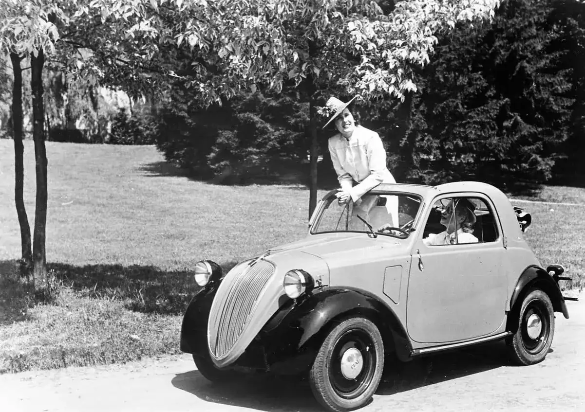 Elegancka kobieta w kapeluszu opiera się o uroczy fiat mini. W środku siedzi druga pasażerka.