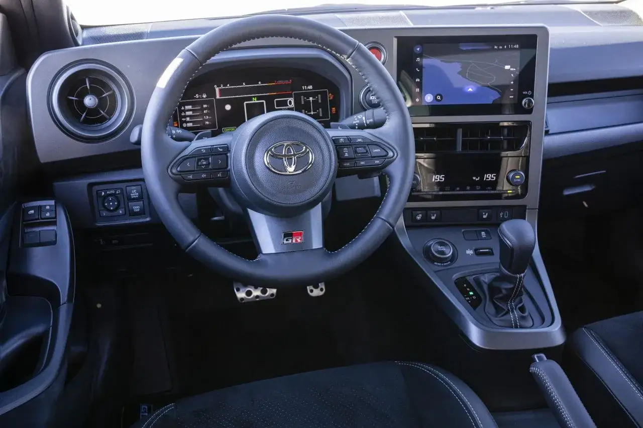 Wnętrze Toyota Yaris GR Sport: kierownica z logo GR, cyfrowy kokpit, ekran multimediów z nawigacją i automatyczna skrzynia biegów.