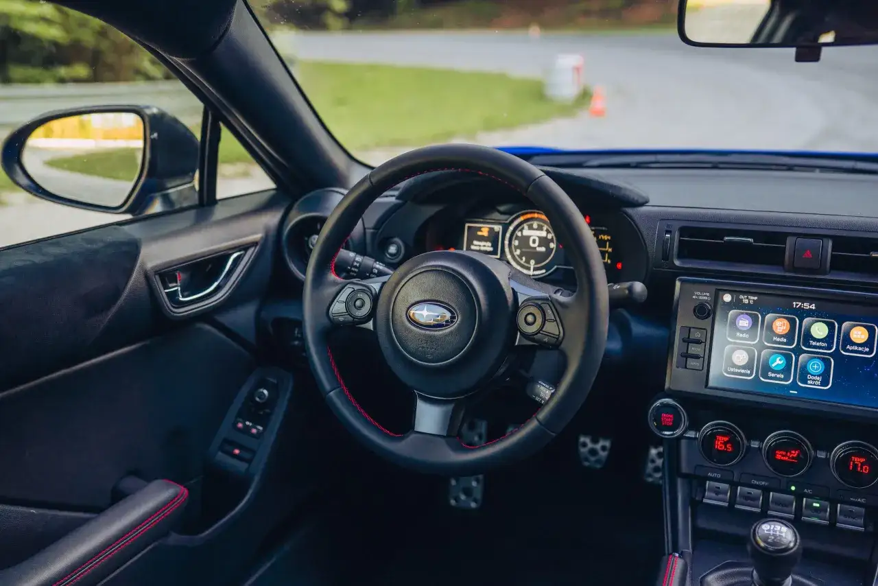 Wnętrze sportowego auta, kierownica z czerwonymi przeszyciami, ekran multimedialny i dźwignia zmiany biegów. To wnętrze toyoty brz.