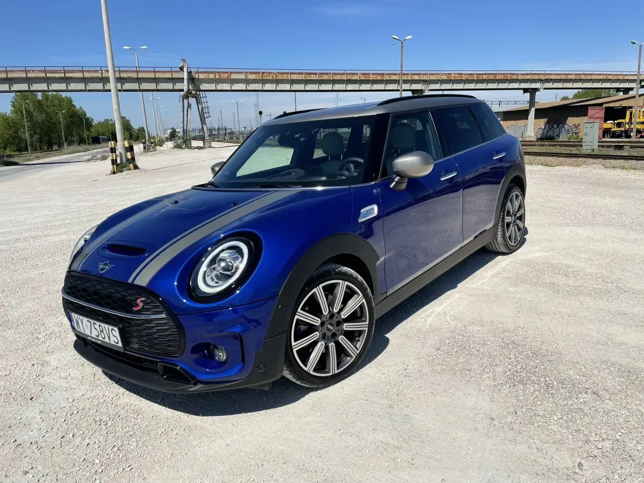 Niebieski mini f56 z szarymi pasami na masce, zaparkowany na żwirowym placu pod wiaduktem.