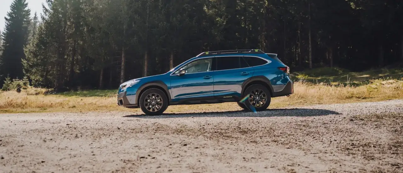 Niebieski Subaru Outback Wilderness gotowy na przygodę w dziczy, zaparkowany na szutrowej drodze wśród drzew.