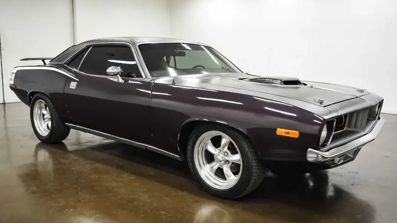 Klasyczny Plymouth Barracuda w ciemnym kolorze, z chromowanymi felgami i sportowym wlotem powietrza na masce.