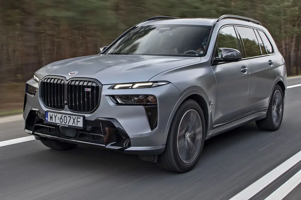 Szary BMW X7 M z czarnym grillem i felgami, jadący po drodze. Sprawdź aktualną bmw x7 cena.