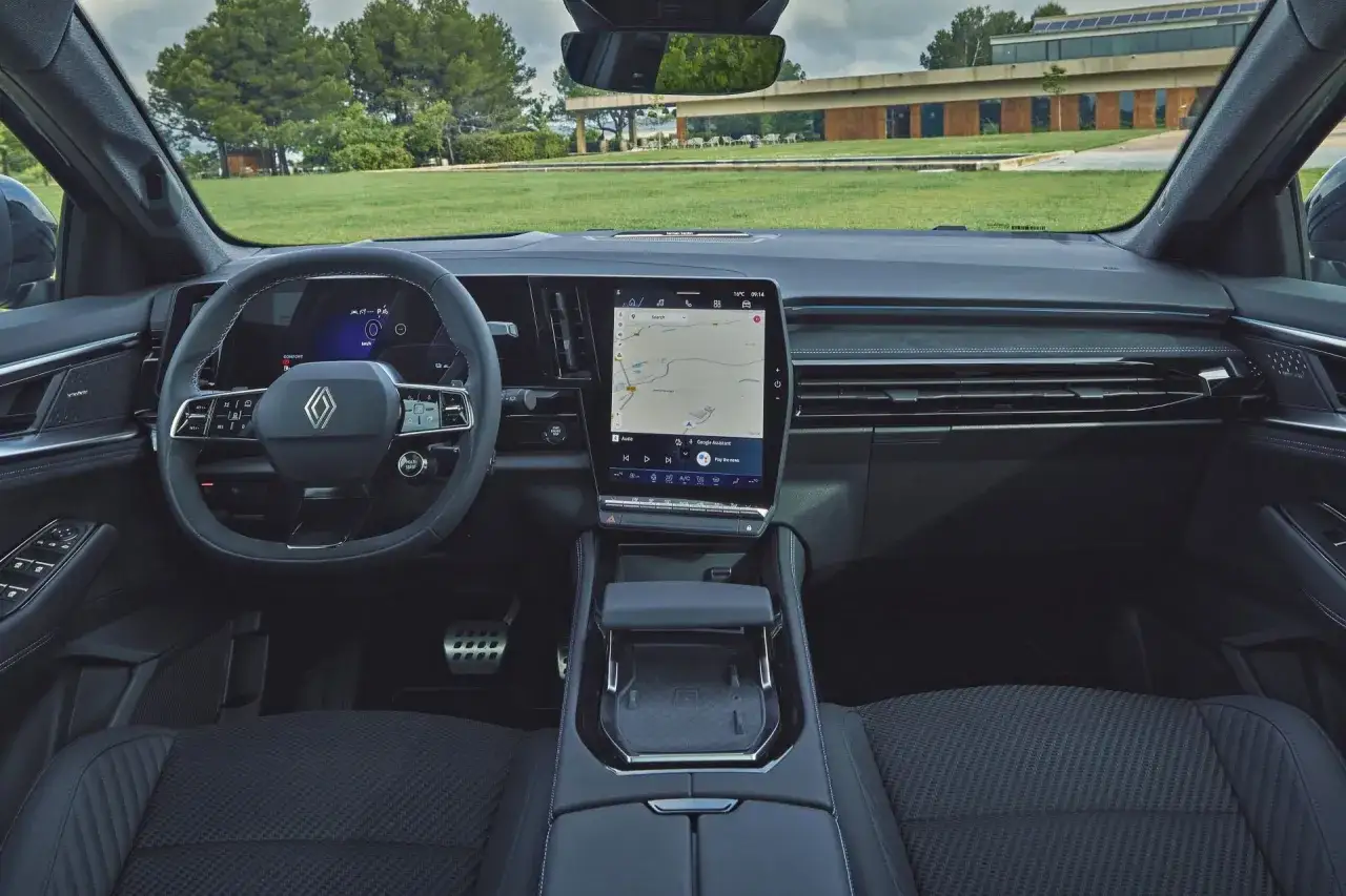 Wnętrze nowego Renault Austral z dużym ekranem multimedialnym i cyfrowymi zegarami. Widok na zielony trawnik i budynek z zewnątrz.
